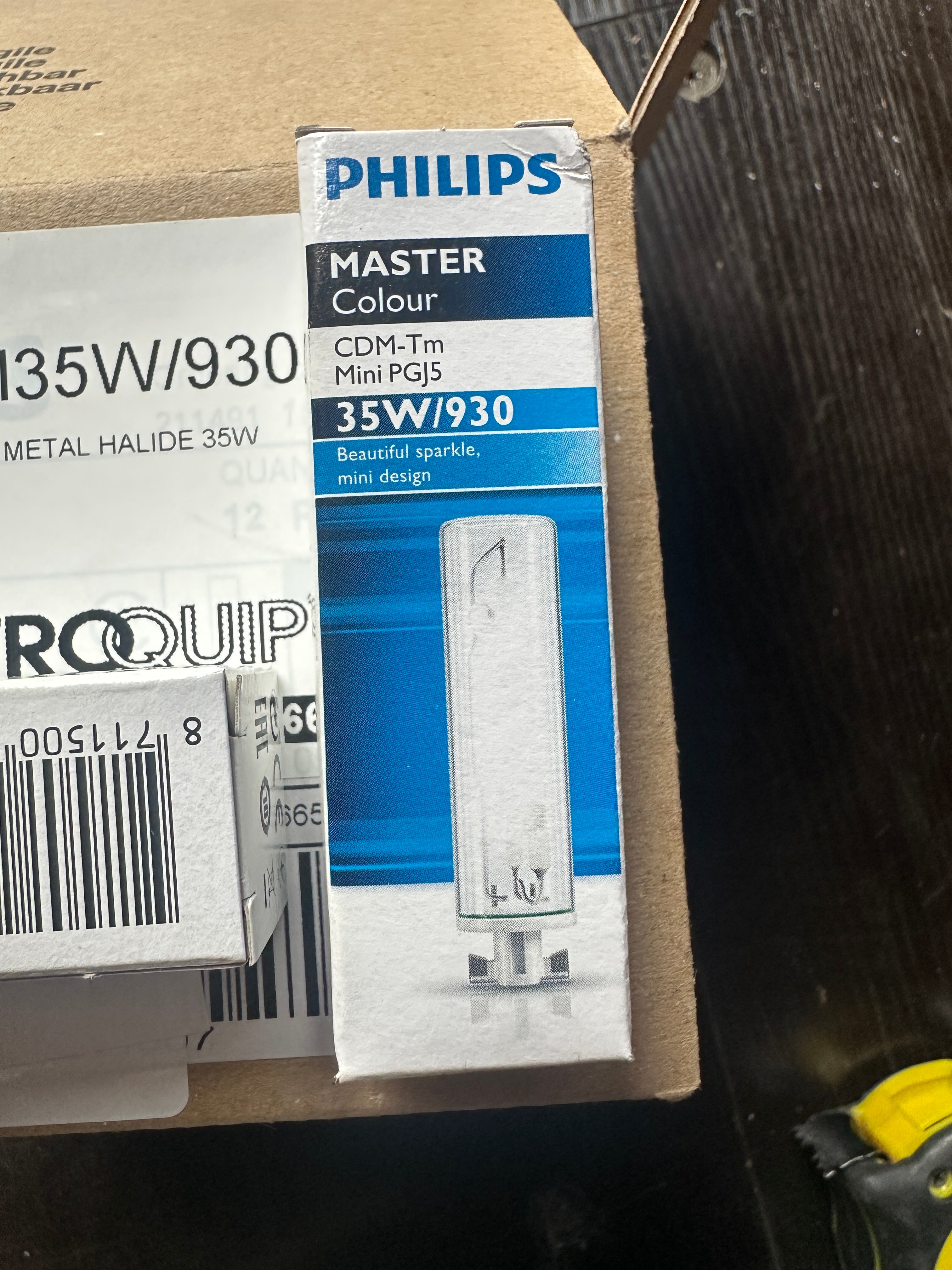 A Philips Master Colour 35W/930 metal halide light bulb box placed on top of another Roquip box on a cardboard shipping box, set on a dark wooden surface.